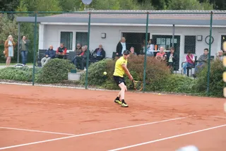 Ein Spieler läuft zum Ball, dahinter Zuschauende vor dem Vereinsheim